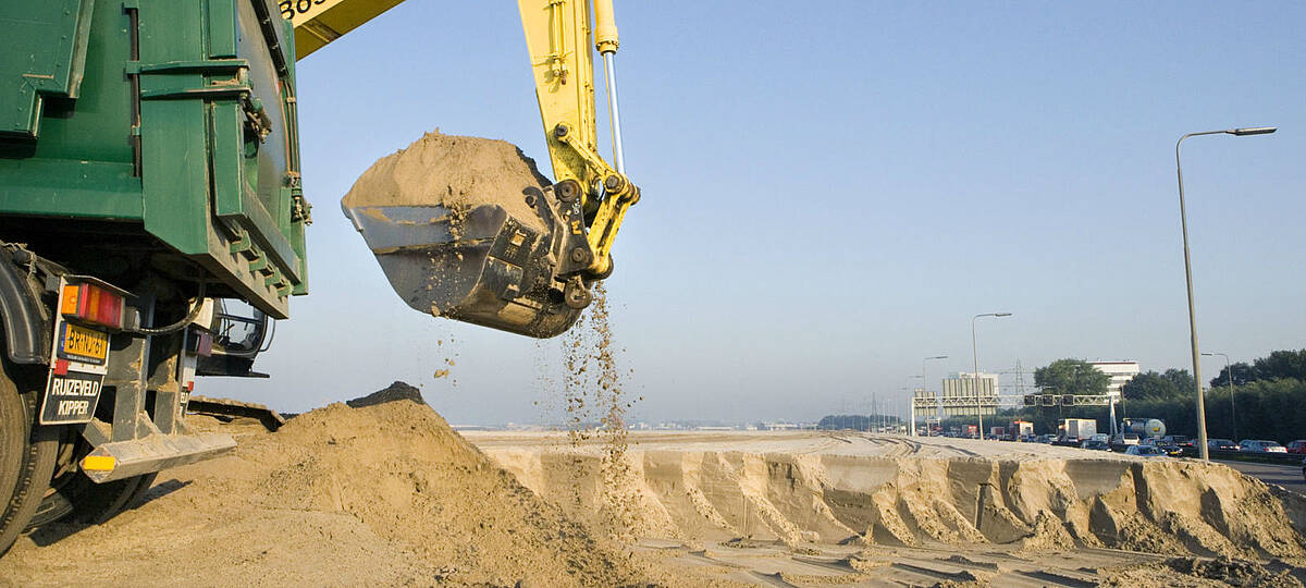 een graafmachine neemt een hap zand om in een vrachtwagen te laden, bij werkzaamheden langs de snelweg.